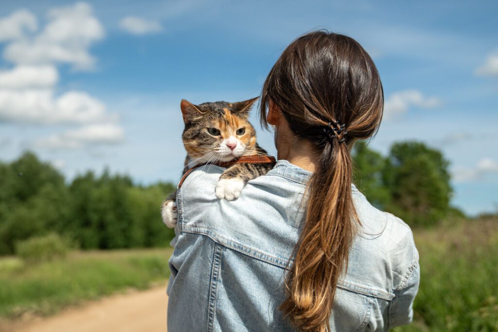 dark haired woman facing away from the camera in a grassy area, holding a calico cat over her shoulder